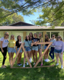 Ten young women stand outside on grass in front of a house, posing with large wooden Greek letters arranged in a triangle shape—a vibrant display of unity among student organizations.
